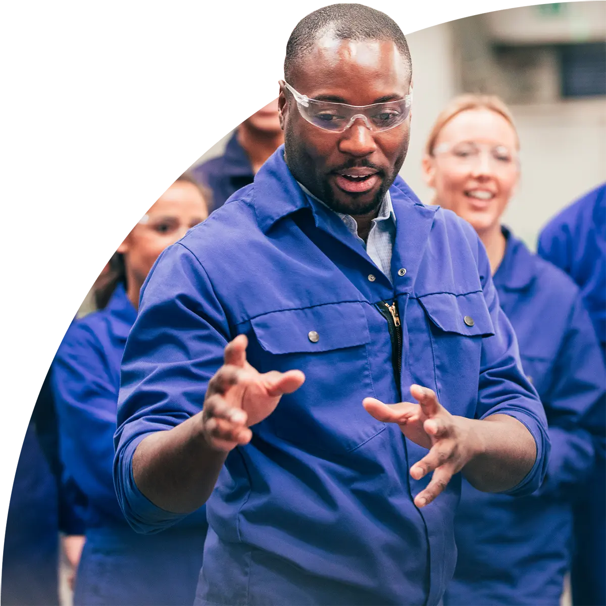 A man in blue coveralls and safety goggles speaks and gestures during a technical training session with colleagues in similar attire.